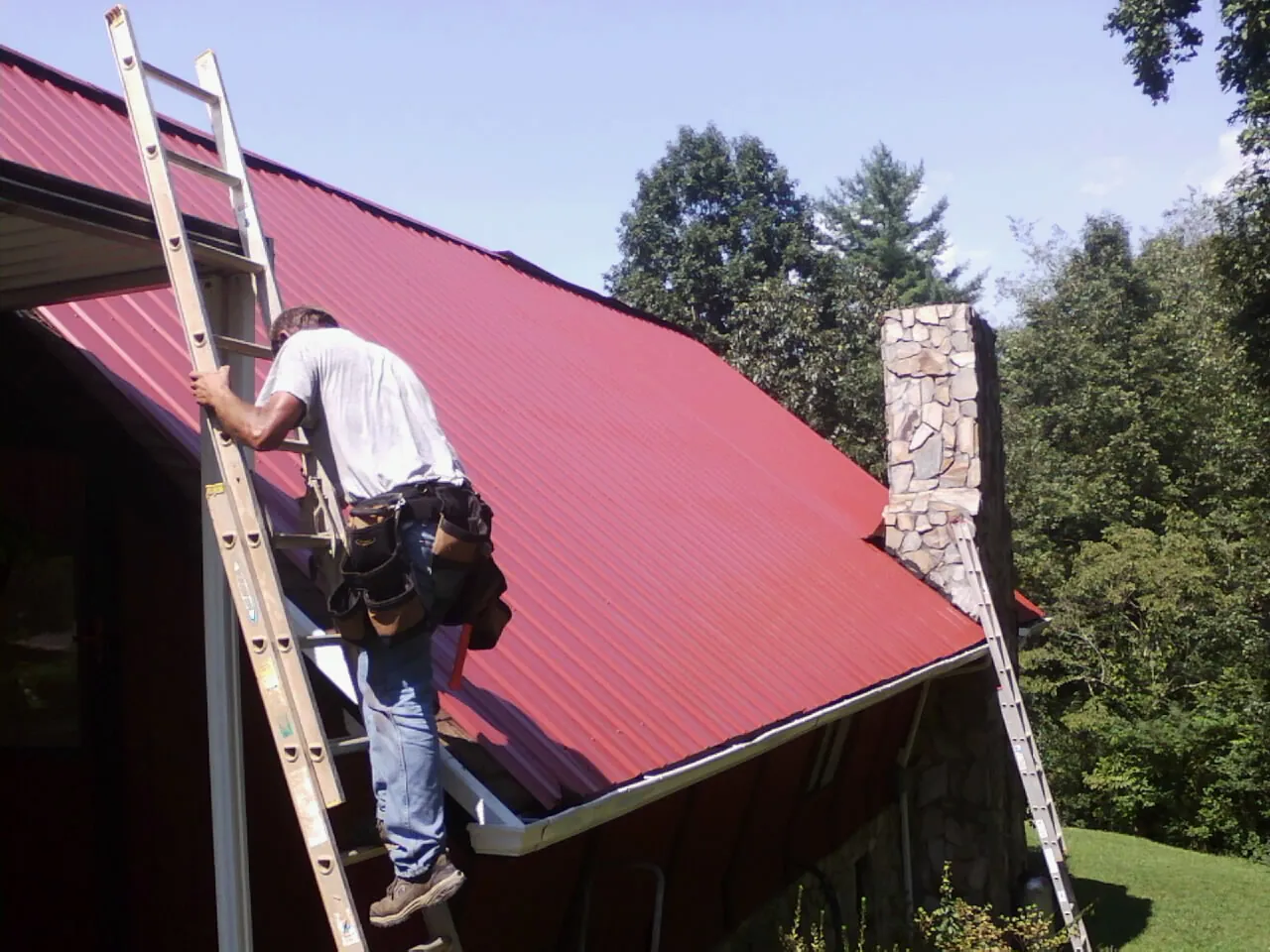 Roofer on a ladder beside a red metal roof and stone chimney — Soffit & Fascia Repair by Roofing Brothers in Monaca